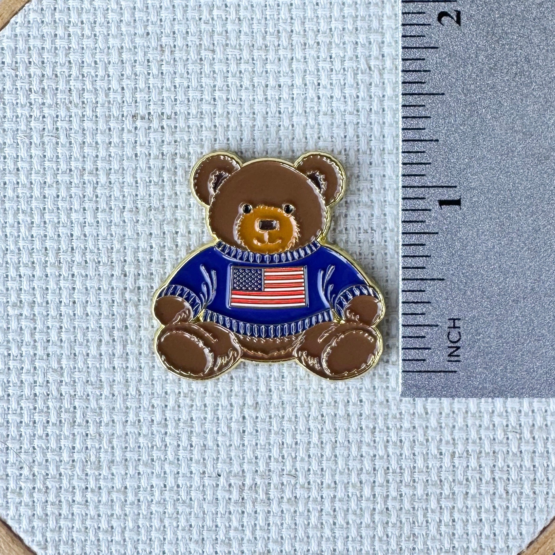Teddy bear enamel needle minder wearing a blue sweater with an American flag, magnetic needle holder for cross stitch, needlepoint, embroidery, quilting, sewing and other stitching projects.